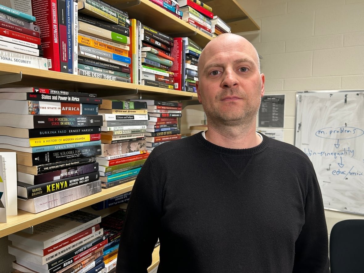 A man wearing a black sweater poses for a photo. Shelves of books are shown behind him.