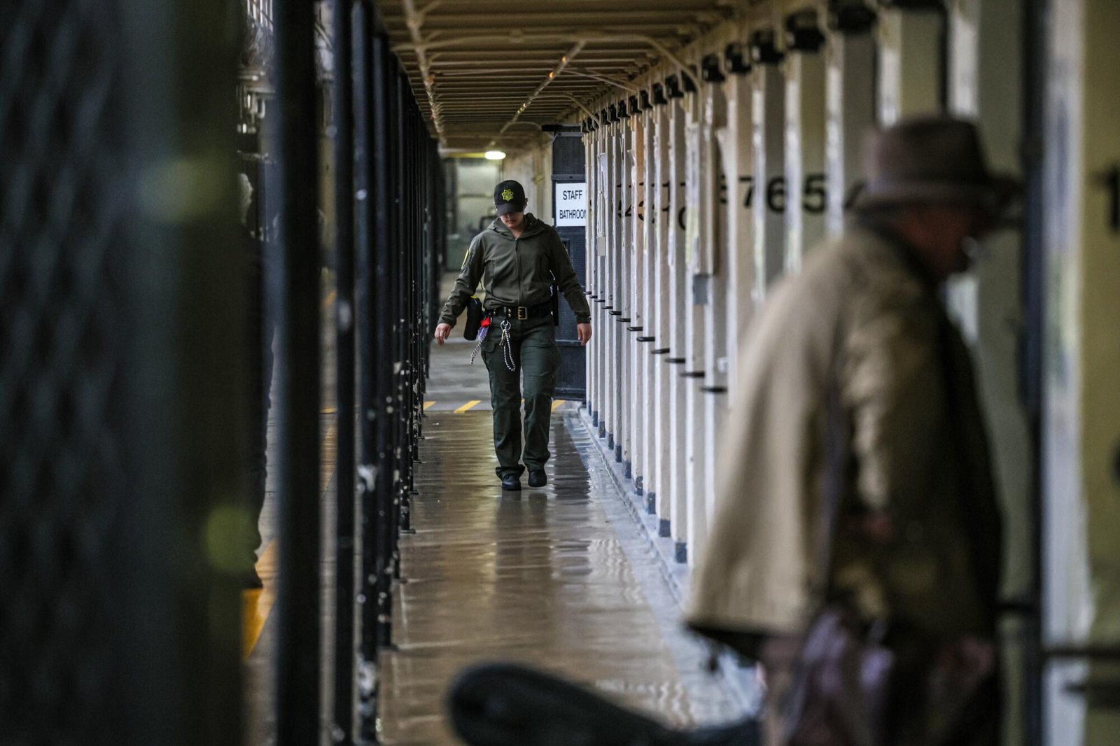 A guard walks along a narrow hallway between prison cells