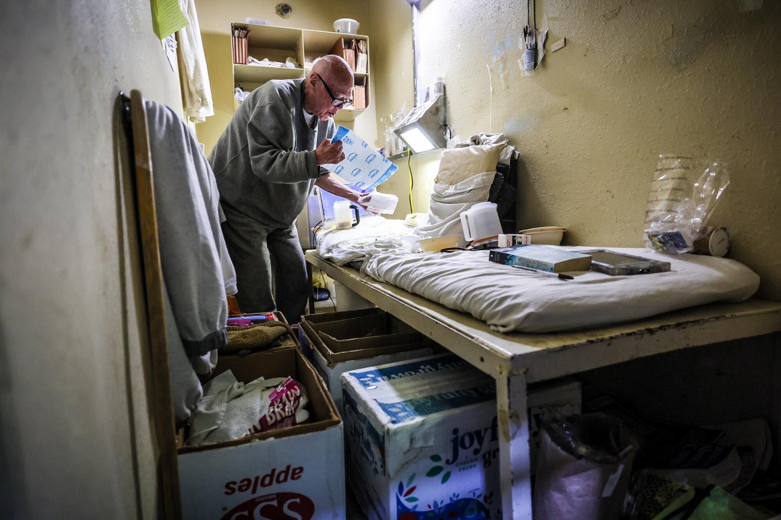 A man with glasses stands near a bed in a cramped prison cell filled with boxes and paperwork.  