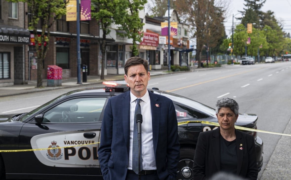 A man stands at a microphone beside a woman; both stand in front of a police car behind caution tape.