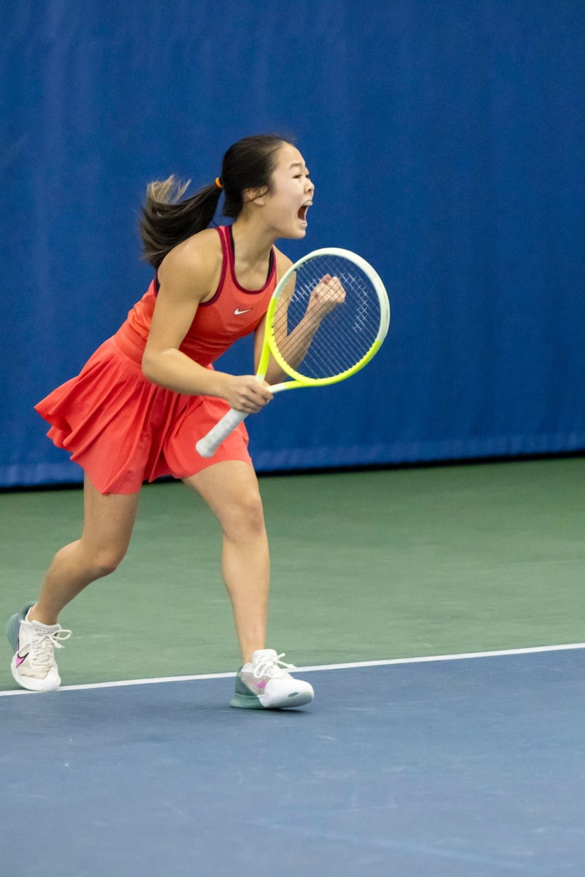 A girl in a red tennis dress holds a racket and shouts with her hands in the air.