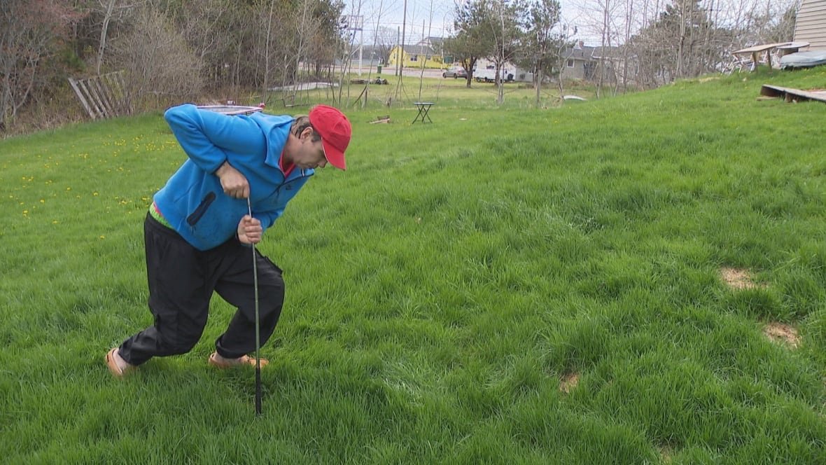 A man is shown halfway up on his feet in a grassy yard, leaning heavily on a golf club.