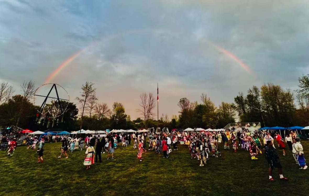 Hundreds of people on green grass against a blue sky with a rainbow in the background and a large circle structure off to the side It's a giant dreamcatcher.