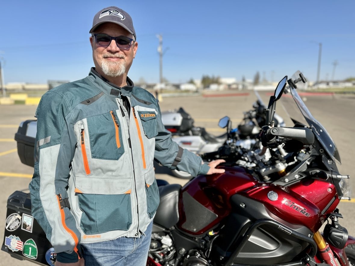 A man in a green coat and sunglass stands next to his motorcycle in an parking lot behind a blue sky. 