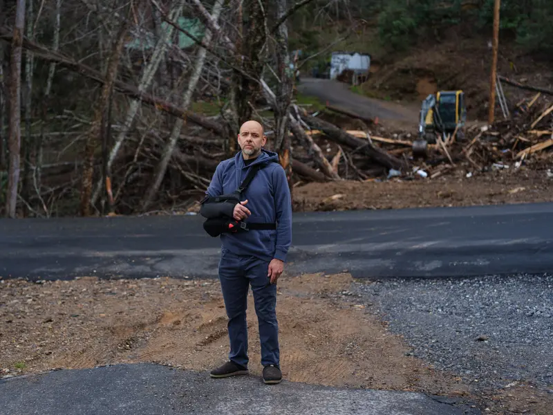 A man with an arm in a sling stands on a road in front of destroyed trees.