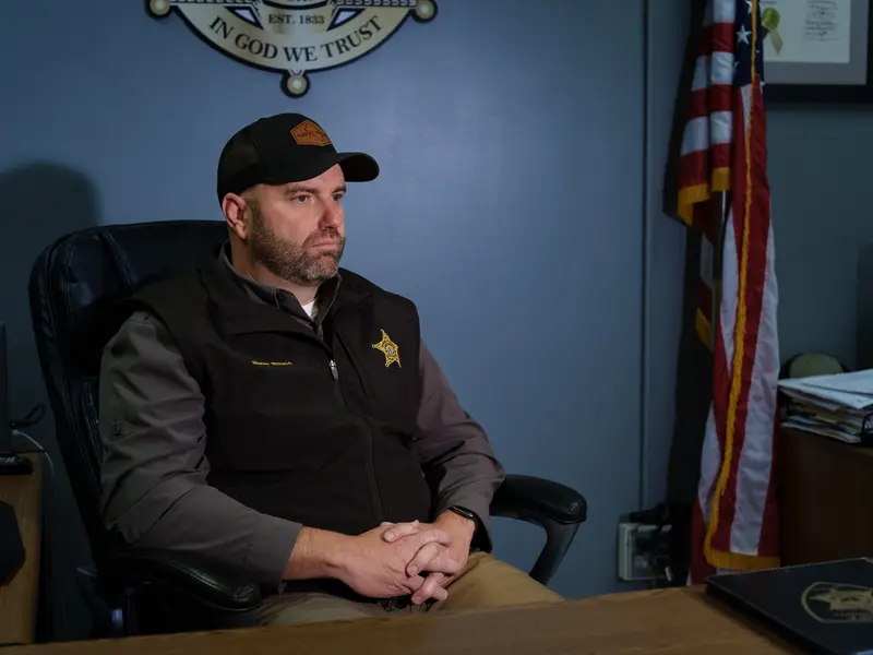 A man wearing a vest with a sheriff’s star sits in a desk chair next to an American flag and a sign with the text “In God We Trust.”