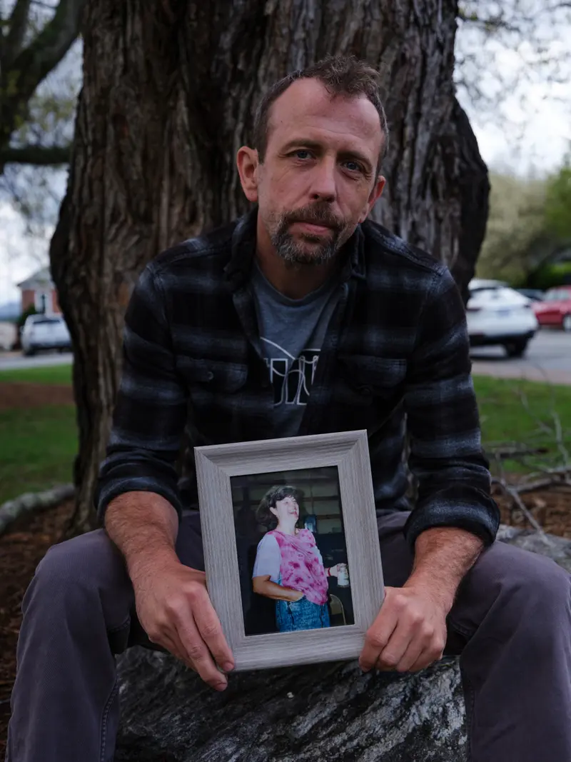 A man sits against a tree trunk holding a framed portrait of a woman laughing.