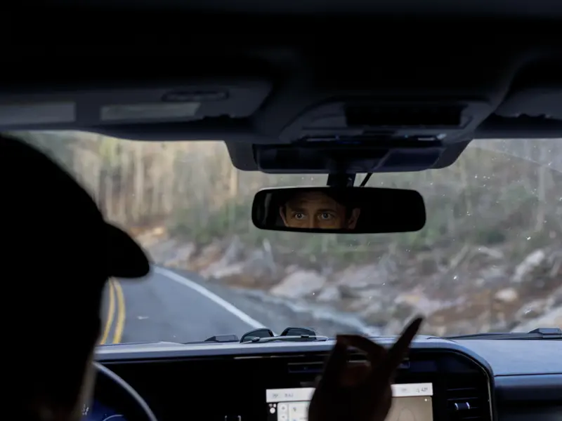 A male driver looks into a car’s rearview mirror as he points at the road outside the windshield.