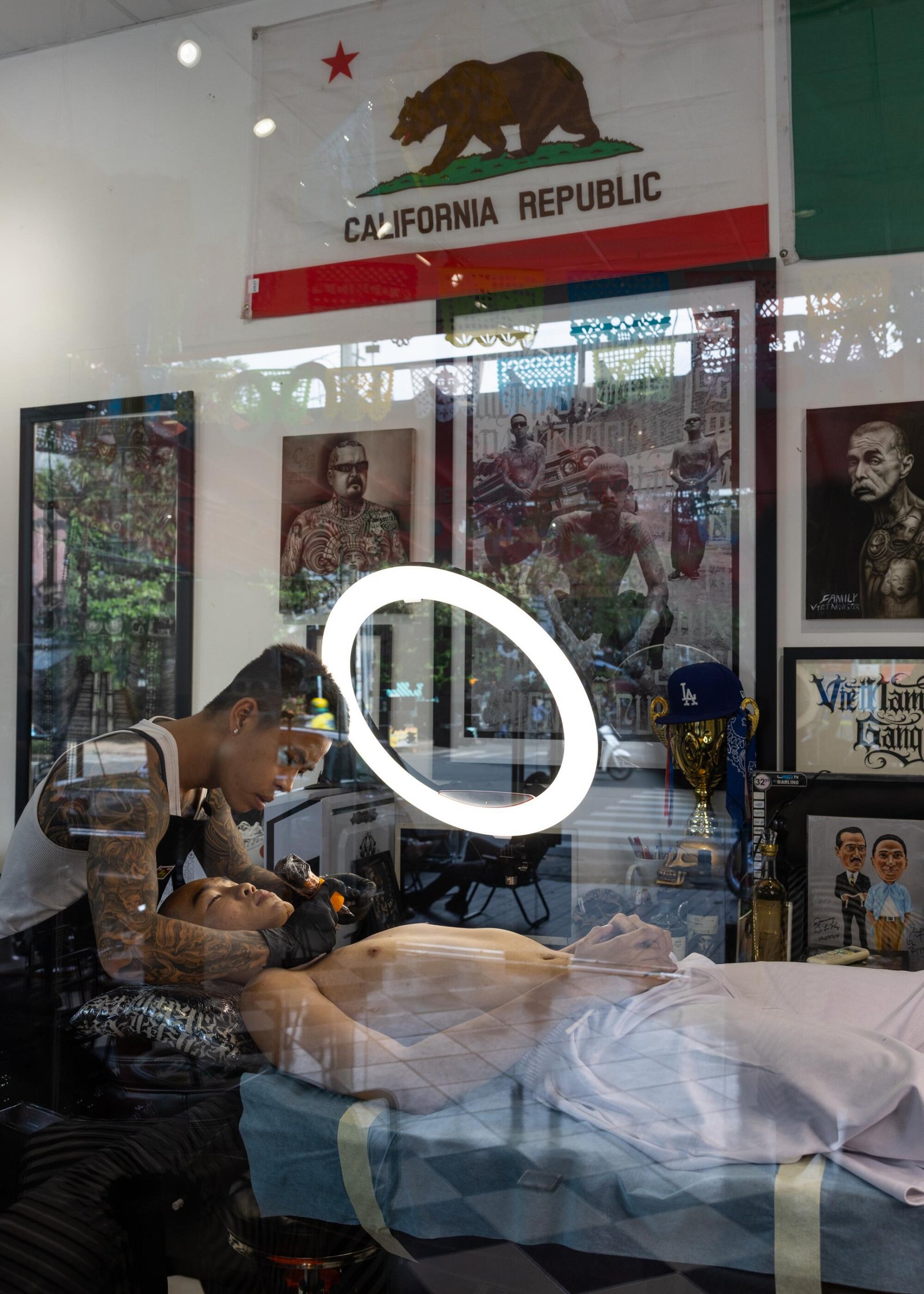 A man giving another man, who is lying down, a tattoo