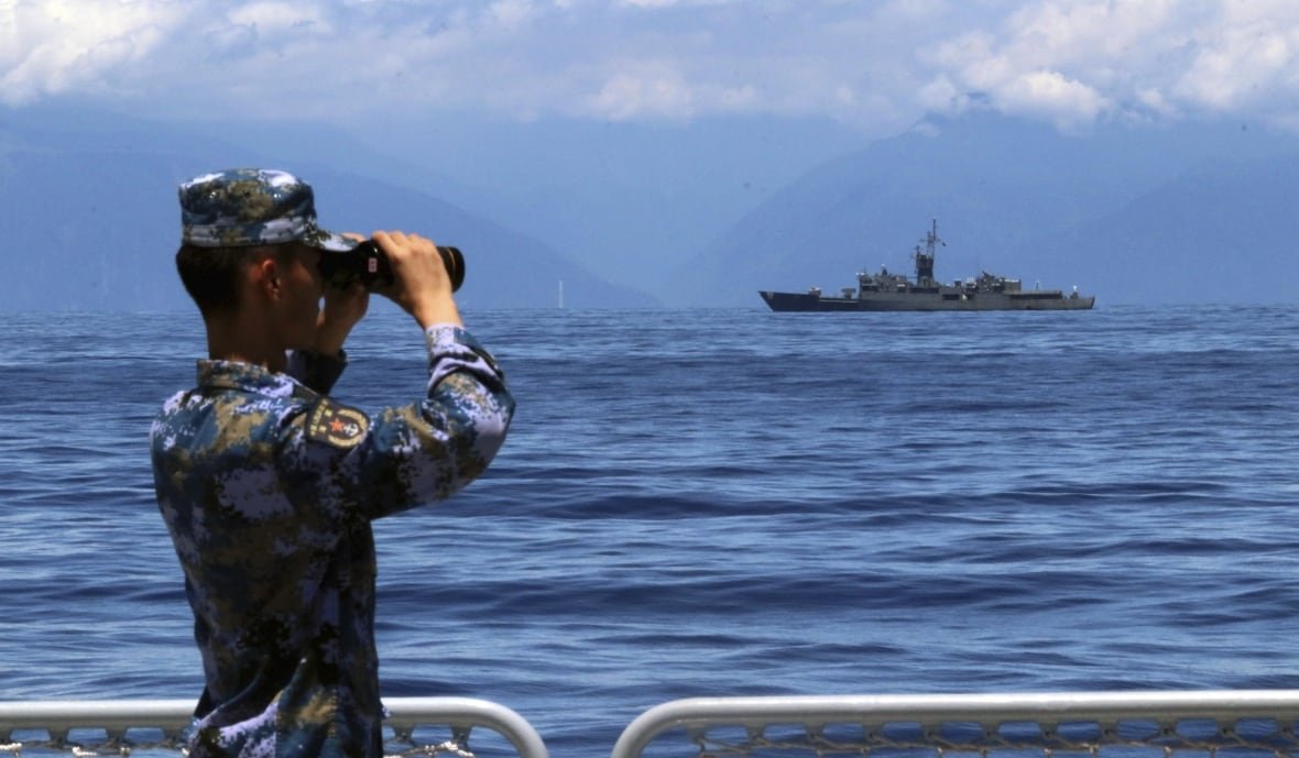 A soldier looks through binoculars at a ship across the water.