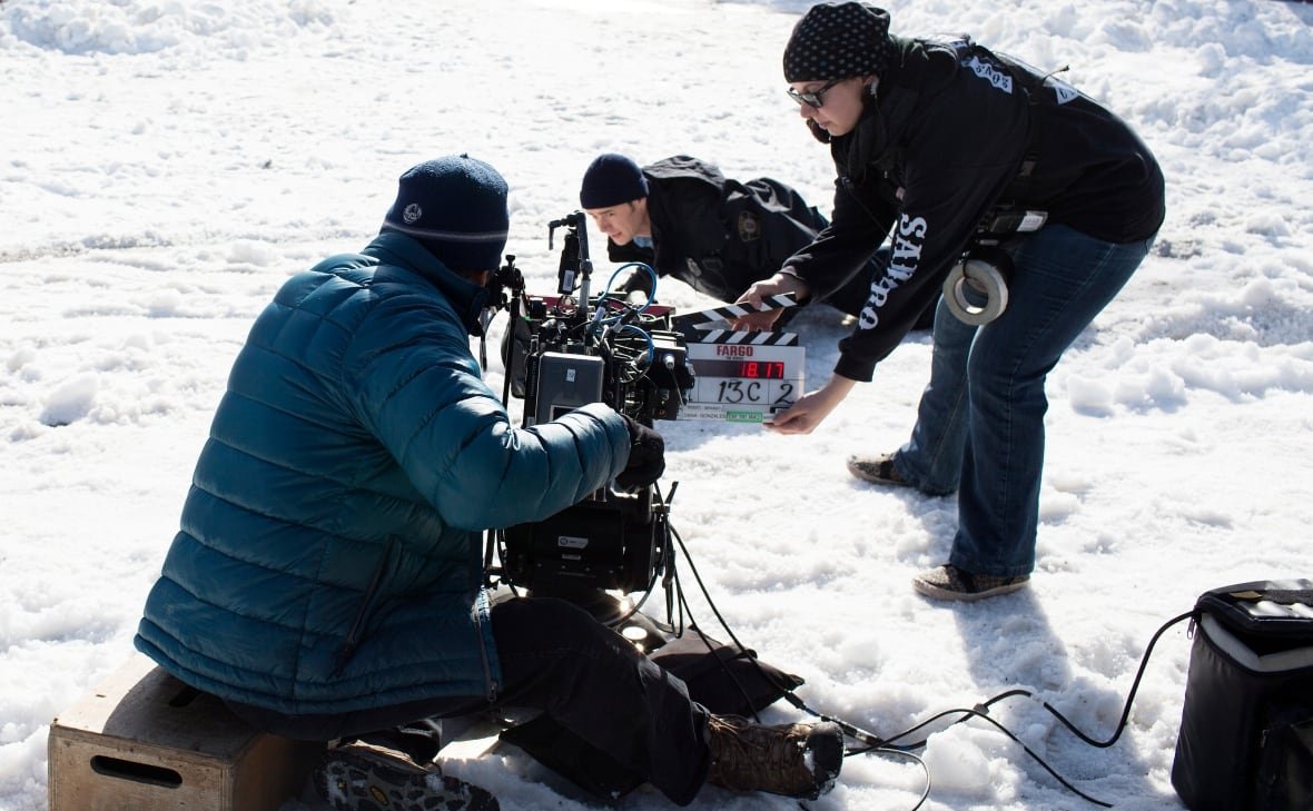 Two members of a film crew crouch in the snow, filming an actor who is on the ground. 