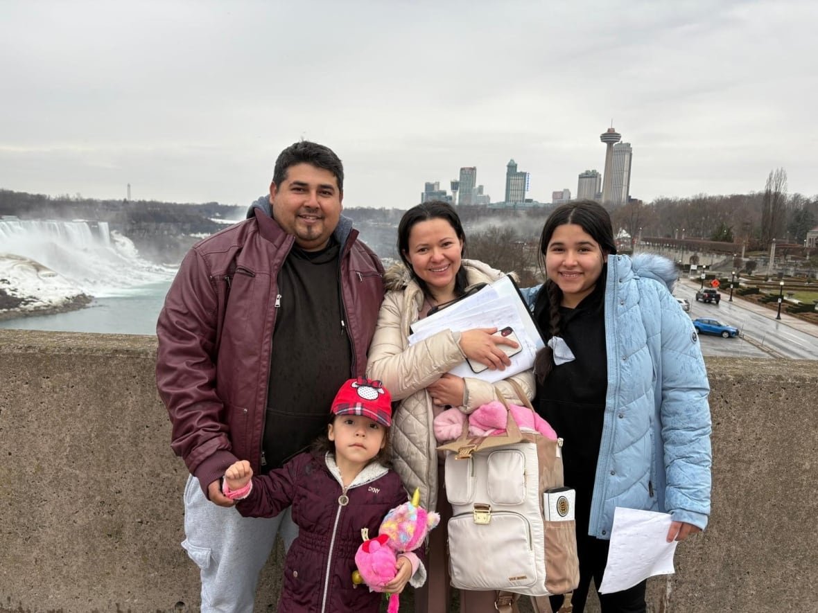 A family of four. The father, on the left, with a young girl between him and the mother, in the centre, and a teenage girl on the right. They are on a bridge in front of Niagara Falls.
