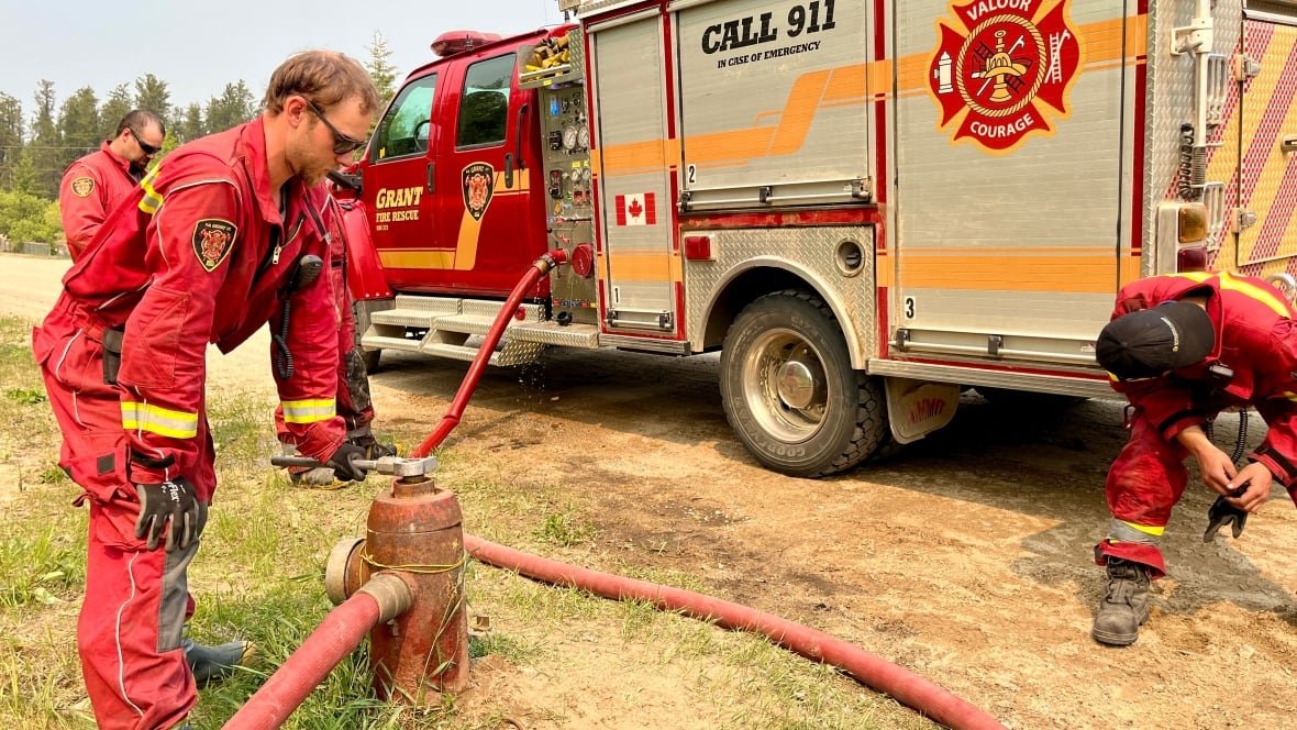 Several men in firefighting gear stand near a truck that has a hose running from it to a hydrant.