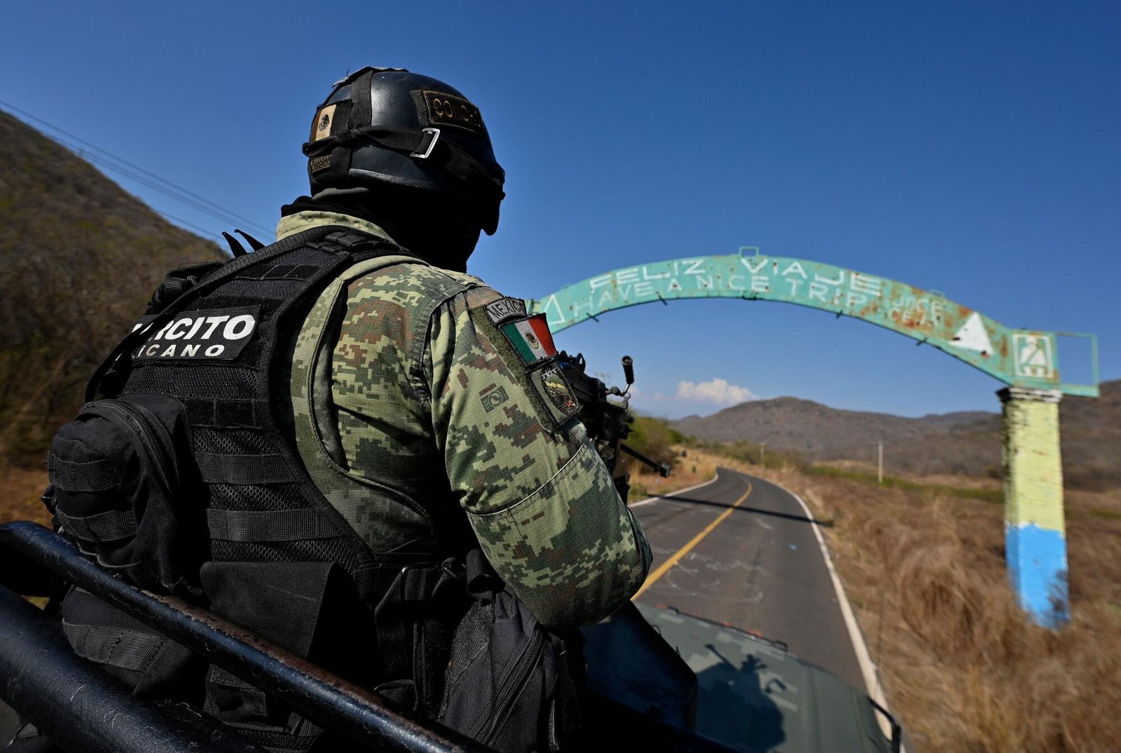 A Mexican soldier in combat gear rides in a patrol vehicle on a rural highway