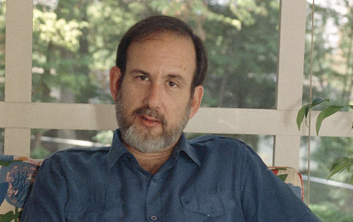 Michael Ledeen on floral sofa in front of plants and a window facing trees in sunshine.