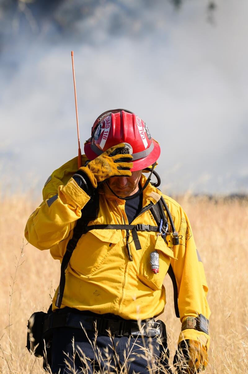  A firefighter in a red helmet walks through a smoky field.  