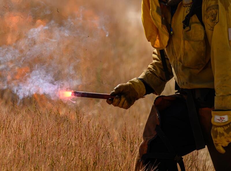 A person in protective gear uses a drip torch to set fire to yellow grass. 
