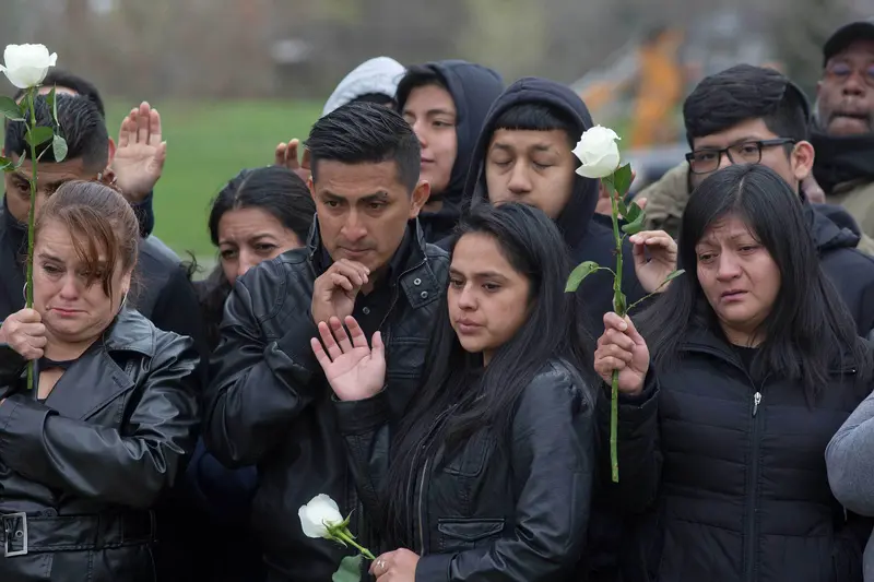 Several people, some of whom are crying, stand close to one another, wearing black jackets and holding white roses.