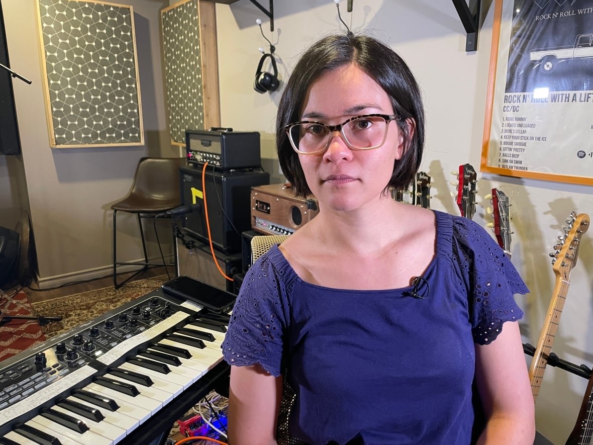 A woman with dark hair, glasses and wearing a blue top sits next to a keyboard in a room of musical instruments.