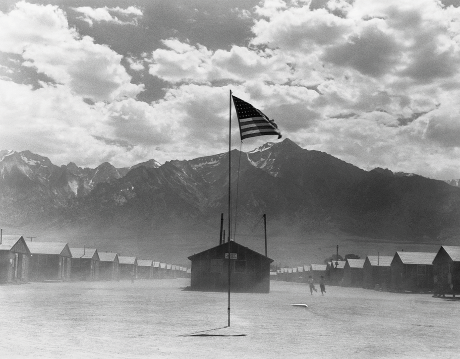 black-and-white photo of dusty camp with rows of barracks on either side and U.S. flag flying from post at center, with mountains in background