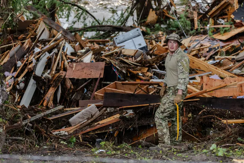 First image: Two men in tank tops, shorts and flip-flops are dwarfed by a fuel tank that has fallen across the road and taken down power lines. Second image: A man in a camo uniform pauses in front of a massive pile of wood, metal and plastic debris.