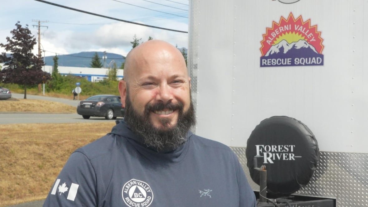 A man with a beard smiles and stands in front of a trailer that says Alberni Valley Rescue Squad.
