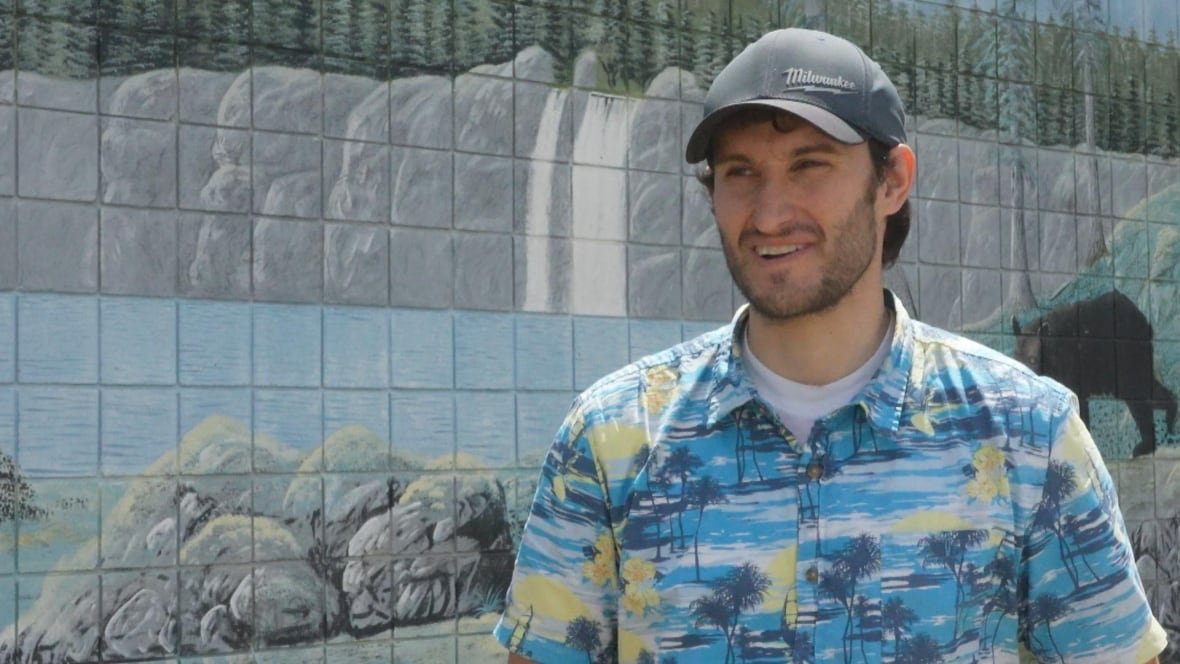 A man wearing a Hawaiian shirt and ball cap smiles in front of a mural.