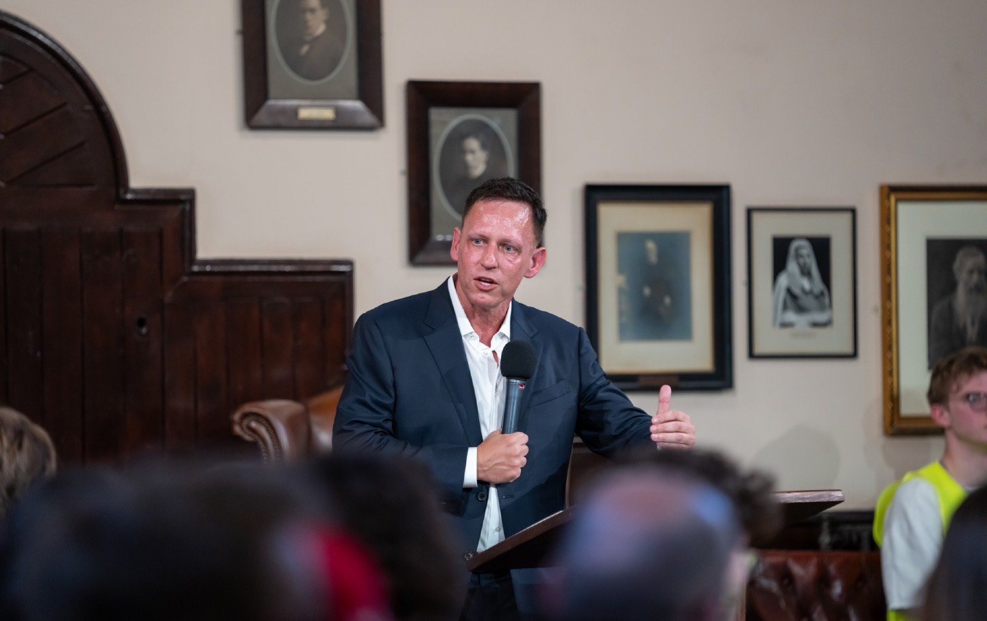 Peter Thiel speaks at a lectern, holding microphone in front of audience at The Cambridge Union.