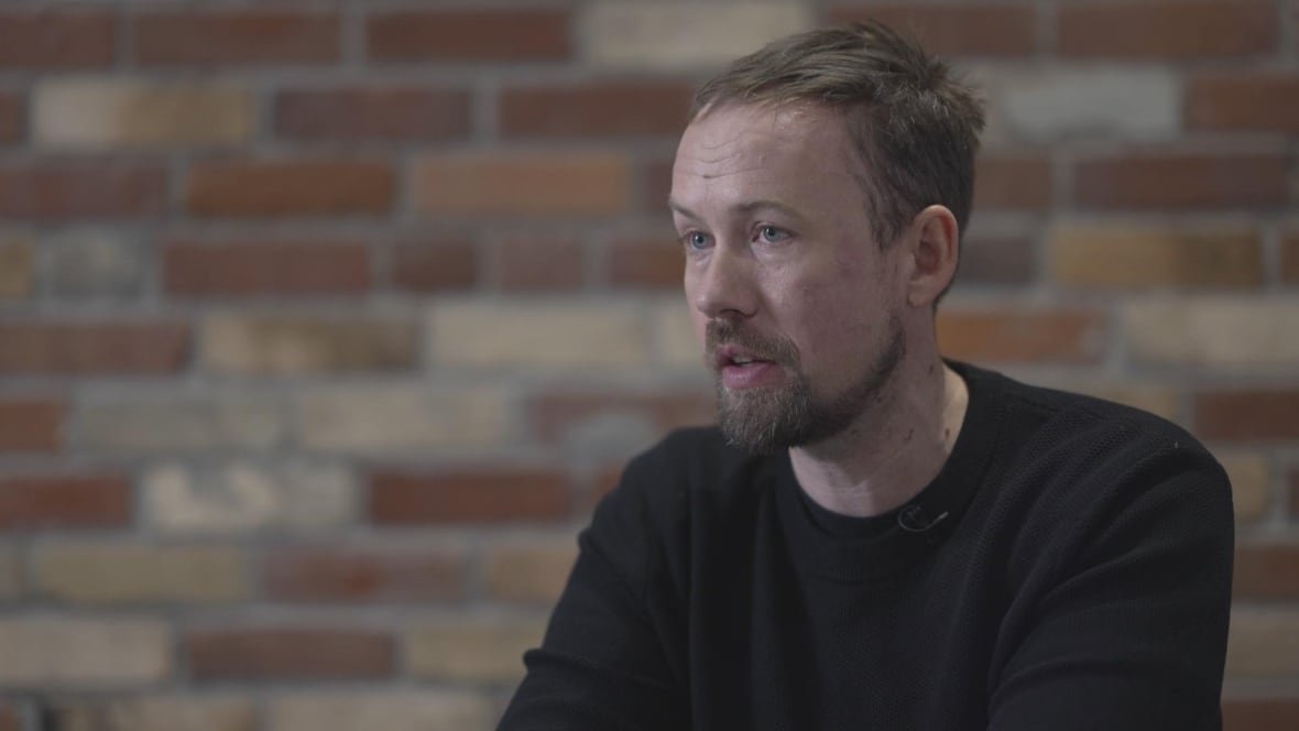 A man in black speaks to the camera.