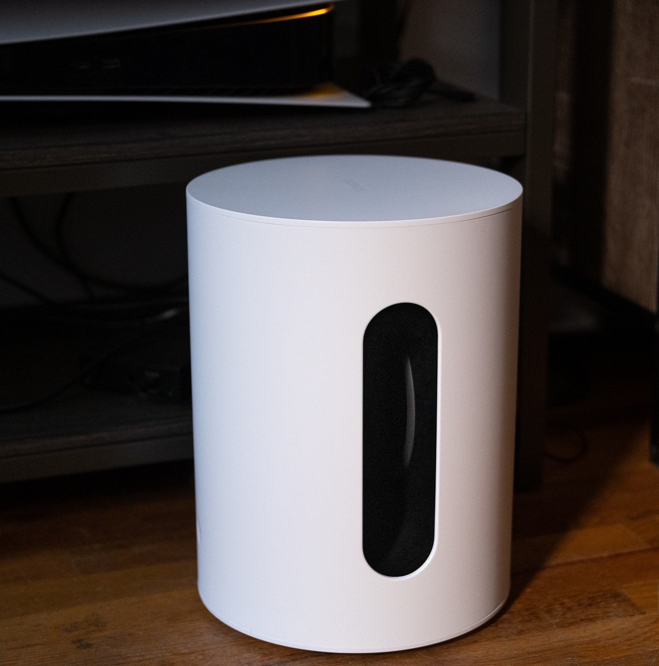 An image of the Sonos Sub Mini pictured on the floor with a TV stand and various components behind it.
