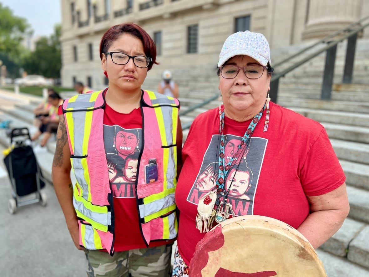 Two people wearing red shirts stand at the foot of steps leading to a large limestone government legislative building.
