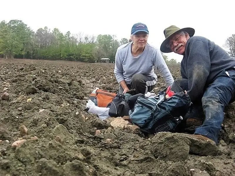 An older couple in muddy clothes pose outdoors on rocky terrain, smiling for the camera.