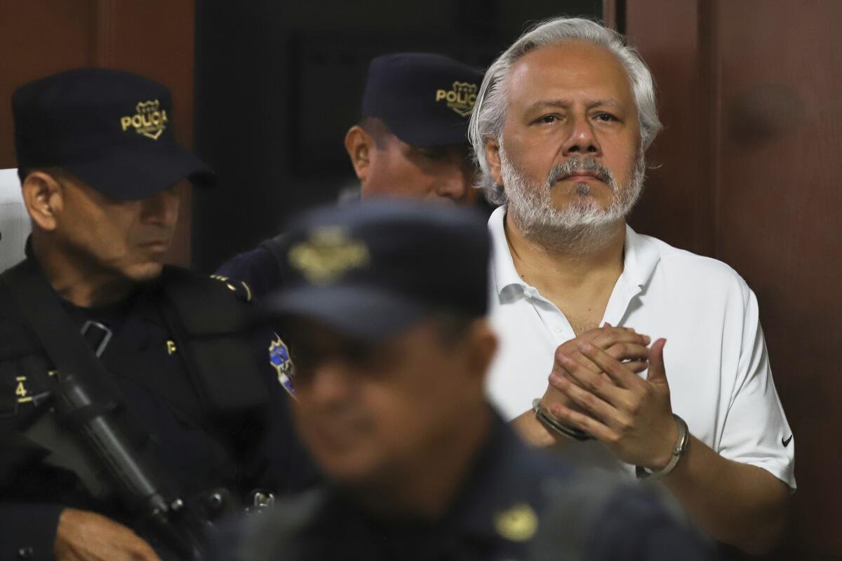 A gray haired man with a gray beard walks, in handcuffs, alongside men in dark uniforms carrying guns.