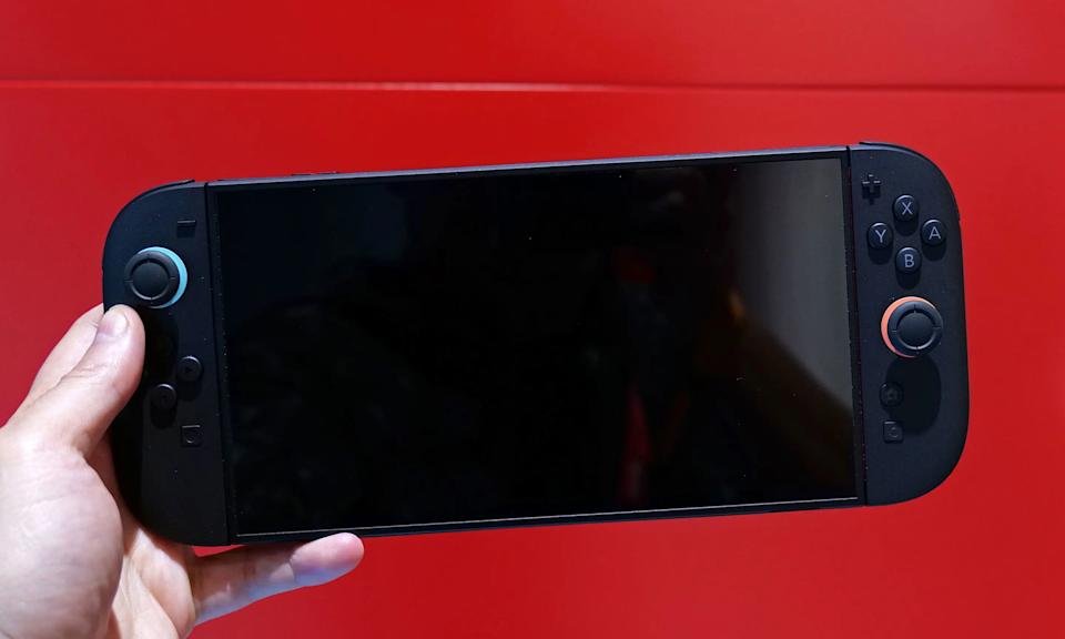 The Nintendo Switch 2 game console is held up with its screen turned off in front of a red wall.
