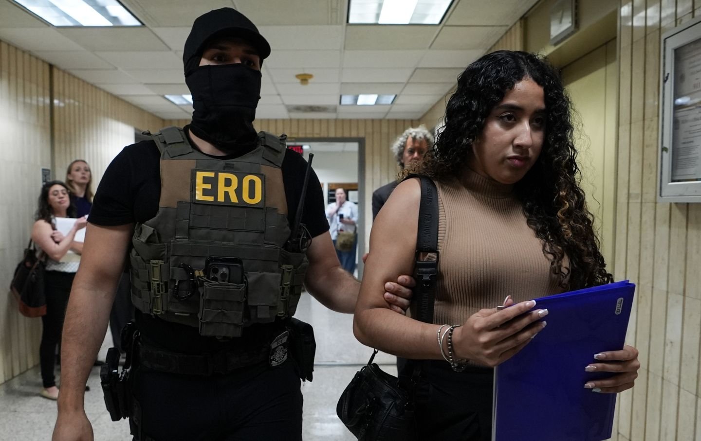 A deportation officer holds a young woman’s arm as they walk down a hall of Federal Plaza courthouse.