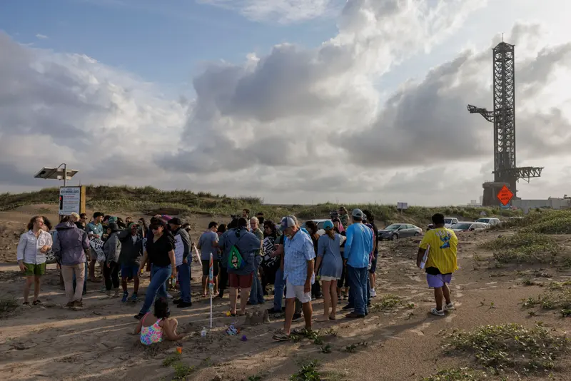 A group of people gathered on a sandy beach with a spaceship launch site looming in the background.