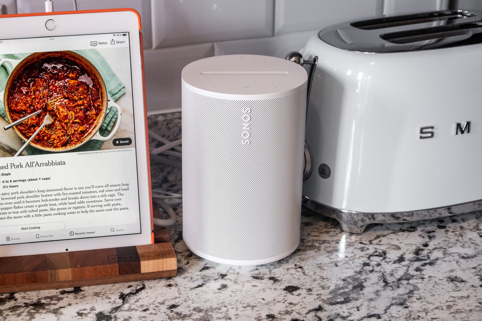 A photo of the Sonos Era 100 speaker in a kitchen setting beside an iPad and toaster.