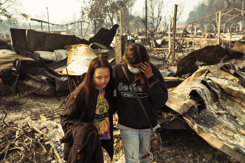Two women hold each other as they walk toward the camera. Burnt homes, trees and structures cover the landscape around them.