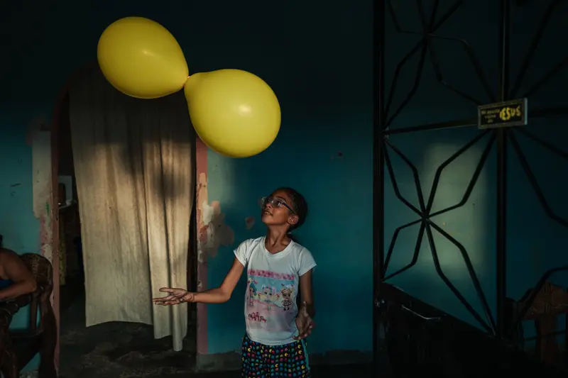 A child looks up at two balloons tied together floating above her. Behind her is a blue wall with some chunks of plaster missing and a doorway covered by a curtain.