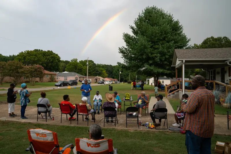 People stand and sit in folding chairs outside, with a rainbow arcing across a cloudy sky.