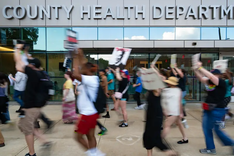 Blurry people march holding signs in front of a glass wall of the County Health Department building.
