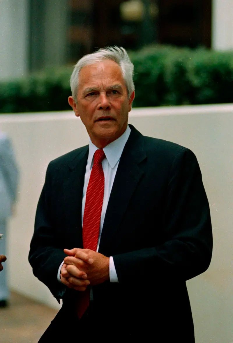 A man with white hair and wearing a blue suit and red tie clasps his hands in front of his body. He is standing outside in front of a white wall and a green hedge.