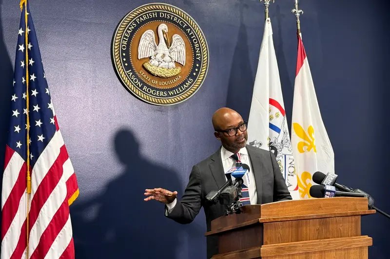 A man wearing a gray suit and black and red striped tie stands at a podium with microphones, in front of the flags of the U.S. and New Orleans.