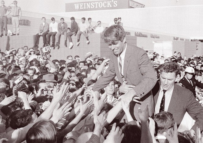 black-and-white archival photo of RFK Sr. smiling and standing above crowd of people with outstretched arms and shaking hands