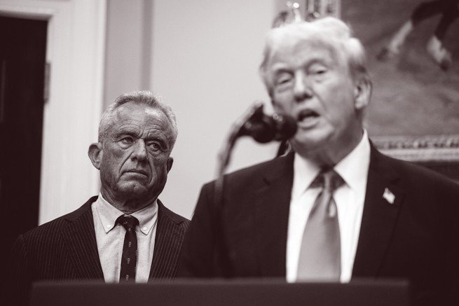 black-and-white photo of Trump speaking into microphone at podium with RFK Jr. looking on from background