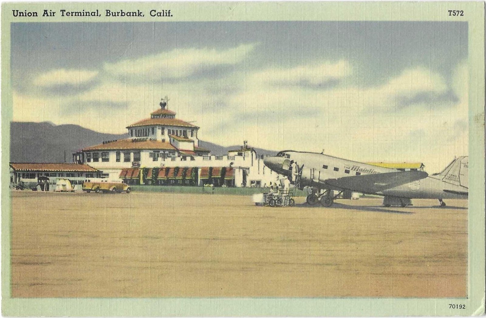A silver plane in the foreground, with the tile-roofed Burbank Airport building in the background.