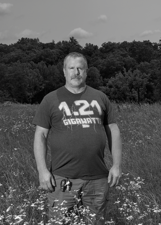 black-and-white photo of man wearing t-shirt with '1.25 Gigawatts' on it and pants standing in field of yellow flowers 